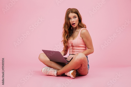 Photo of young amazed readhead woman with laptop, siiting with crossed legs