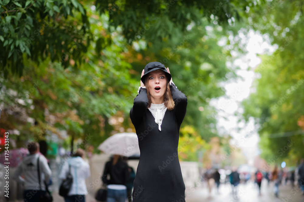 A beautiful thin girl in an amazing black dress in retro style on the background of a pedestrian street. A girl in a black hat after the rain. Emotion of the girl. Emotional face.