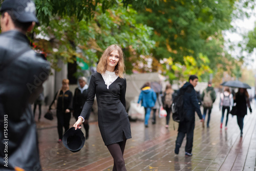 A beautiful thin girl in an amazing black dress in retro style on the background of a pedestrian street. A girl in a black hat after the rain. Emotion of the girl. Emotional face.