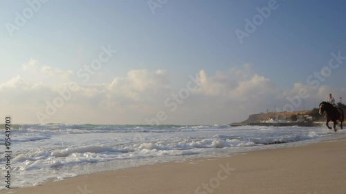 Beautiful young women riding dark horses at sea beach. Enjoying beautiful landscape.Galloping run in sunset or sunrise light and splashing water drops around.Riders running at a gallop.Slow motion.