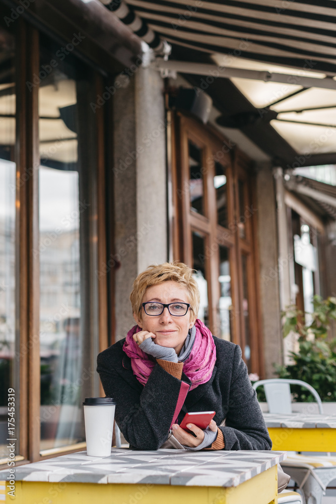 Dreamy woman sitting in outside cafe with coffee and smartphone and looking at camera. Dreamy woman sitting in outside cafe with coffee and smartphone and looking at camera.
