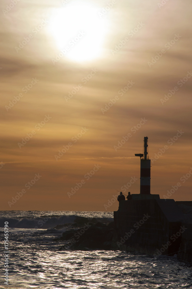 Small lighthouse and fishermen by the beach shore, during sunset