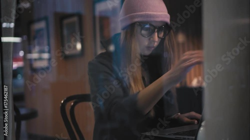 A stylish hipster woman sits down in a cafe for a table, opens the laptop and starts working. View through the window