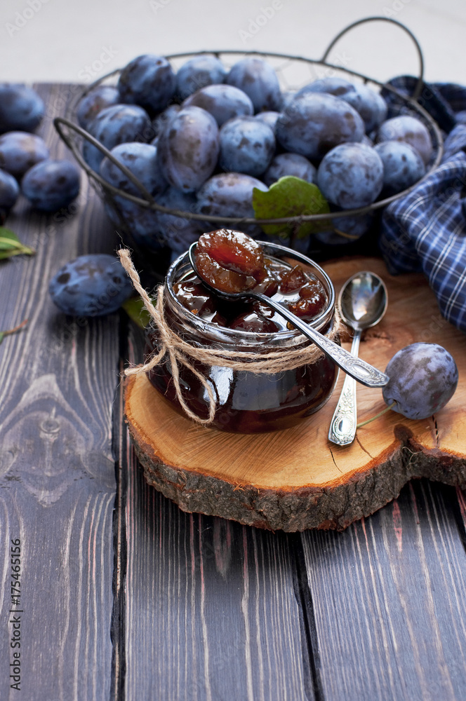 Homemade plum jam in a glass jar and fresh blue plums in a bowl on a dark rustic wooden background top view.