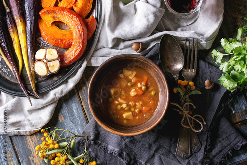 Fall holiday table decoration setting with bowls of hot carrot potato soup, baking pumpkin, carrot, garlic, fresh coriander, red wine, orange berries. Flat lay over wooden table