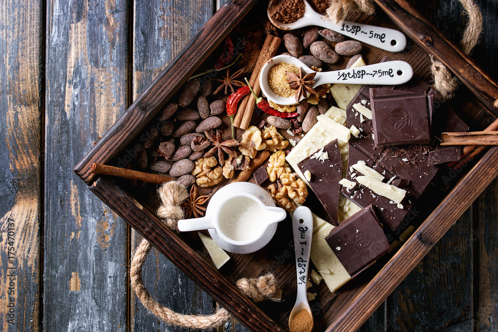 Ingredients for cooking hot chocolate. White and dark chopping chocolate, cocoa powder, cocoa beans, cream, cinnamon, sugar in spoons in wood tray over old wooden background. Top view with space Ingredients for cooking hot chocolate. White and dark chopping chocolate, cocoa powder, cocoa beans, cream, cinnamon, sugar in spoons in wood tray over old wooden background. Top view with space