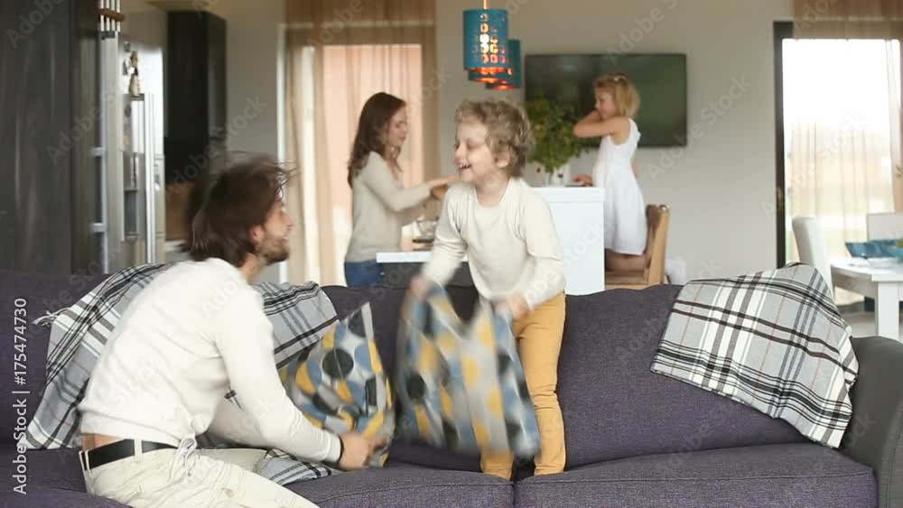Happy family couple spending time with children, having fun with small kids in modern cozy big living room, father playing pillow fight with son on sofa, mother and daughter standing at dining table