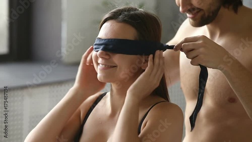 Young man blindfolding smiling woman sitting on bed, couple enjoy foreplay playing erotic role game with bdsm fetish black ribbon on eyes, husband embracing kissing wife before making love having sex