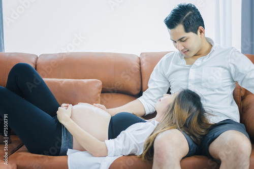 Handsome husband and his beautiful pregnant wife are relaxing together on sofa in the living room, wife is lying on her husband's leg