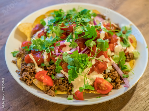 White plate of yellow corn tortilla chips with beef and red onion, cucumber, tomatoes, sauce, salsa and fresh coriander.