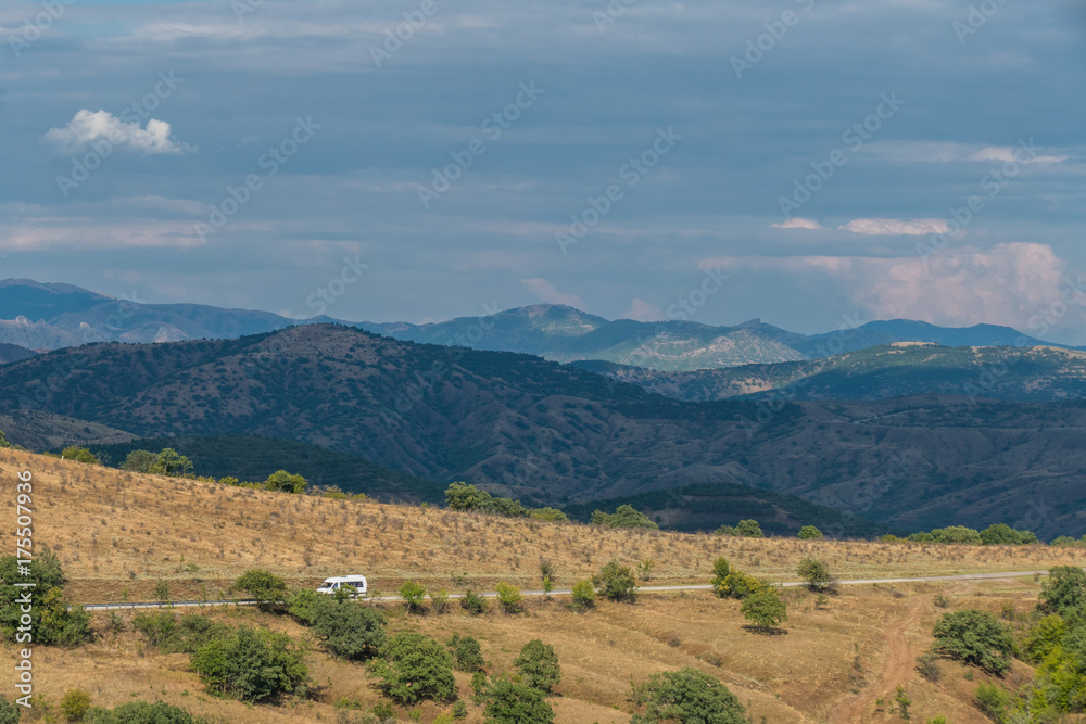 Country road through hilly terrain / Road through hilly terrain on background of blue mountains and gray clouds