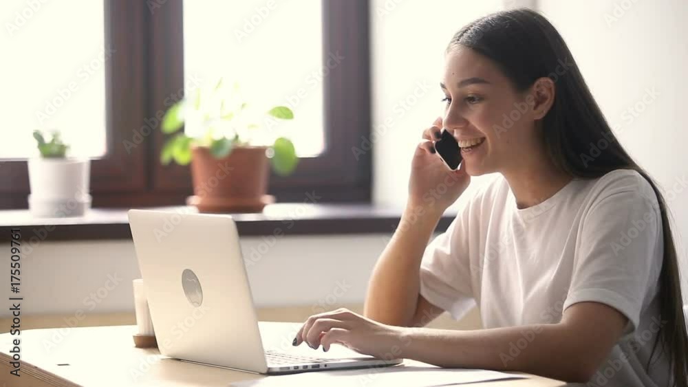 Young casual woman sitting at desk using laptop, answering call, talking on the phone, smiling teen speaking on mobile, having pleasant conversation on cell while working with computer in office Young casual woman sitting at desk using laptop, answering call, talking on the phone, smiling teen speaking on mobile, having pleasant conversation on cell while working with computer in office