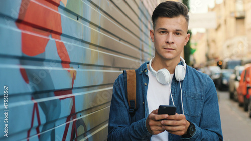 Portrait of attractive young man with head phone on the neck using cell phone for chatting with friends while walking on the street. View from the side.