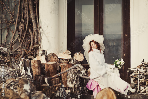 Beautiful ginger bride in a white fur coat with a bouquet of white flowers sits on a stump on a wood background. Winter wedding outdoors.