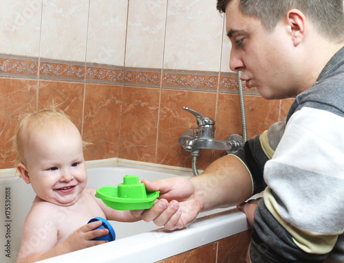 Happy laughing baby taking a bath playing with foam bubbles. Little child in a bathtub. Smiling kid in bathroom. Dad bathes the child. Infant washing and bathing. Hygiene and care for young children.