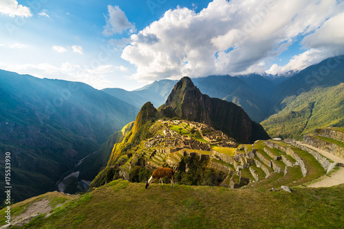 Machu Picchu illuminated by the warm sunset light. Wide angle view from the terraces above with scenic sky and sun burst. Dreamlike travel destination, world wonder. Cusco Region, Peru. photo