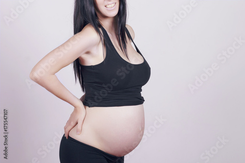 Brunette pregnant woman, with a strong pain, massaging her back ache. Indoors, over a grey wall.