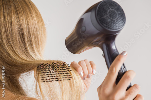 Woman using hair dryer and comb on her blonde hair on the gray background. Cares about a healthy and clean hair. Beauty salon concept.