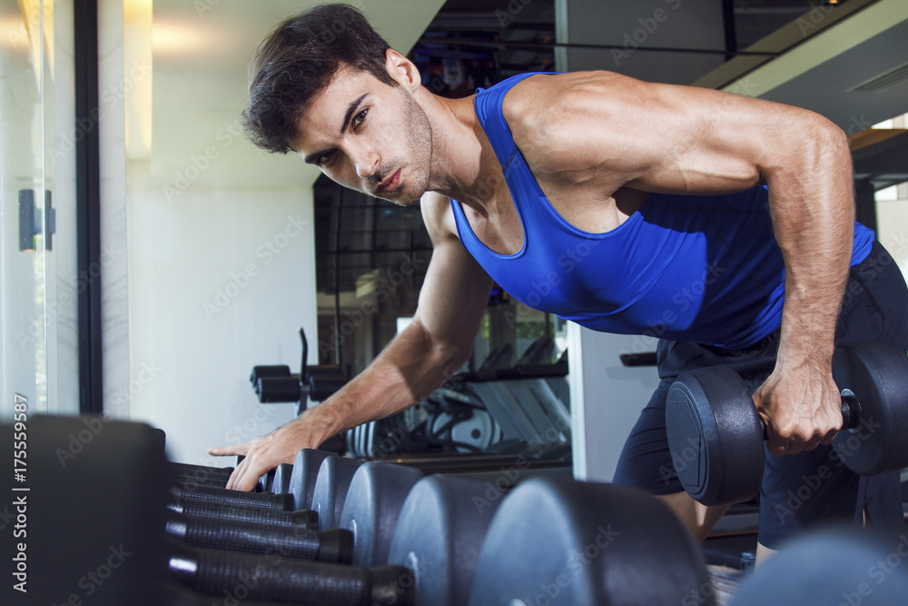 Young handsome muscular man exercising in the gym. One-arm bent-over dumbbell row next to the weight rack. 