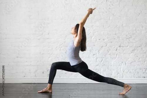 Young attractive woman practicing yoga, standing in Warrior one exercise, Virabhadrasana I pose, working out, wearing sportswear, gray tank top, black pants, indoor full length, studio background
