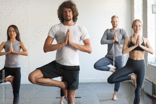 Group of young sporty people practicing yoga lesson with instructor, standing in Vrksasana exercise, Tree pose, working out, indoor close up photo, studio. Wellbeing, wellness concept