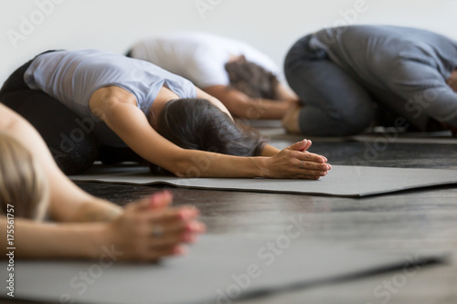 Group of young sporty people practicing yoga lesson with instructor, sitting in Balasana exercise, Child pose, friends working out in club, indoor close up image, studio. Wellbeing, wellness concept photo