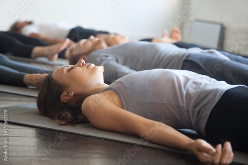 Group of young sporty people practicing yoga lesson with instructor in gym, lying in Dead Body exercise, doing Savasana, Corpse pose, friends relaxing after working out in sport club, studio image