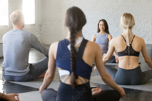 Group of young sporty people practicing yoga lesson with instructor, sitting in Sukhasana exercise, doing Easy Seat pose, working out indoor, studio, rear view. Wellbeing, wellness concept
