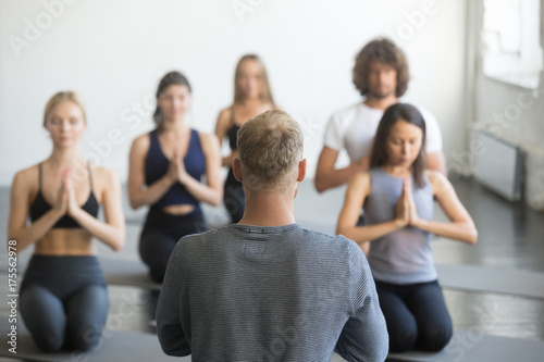 Group of young sporty people practicing yoga lesson with instructor, sitting in vajrasana exercise, seiza pose, working out indoor, studio, rear view at the teacher. Wellbeing and wellness concept