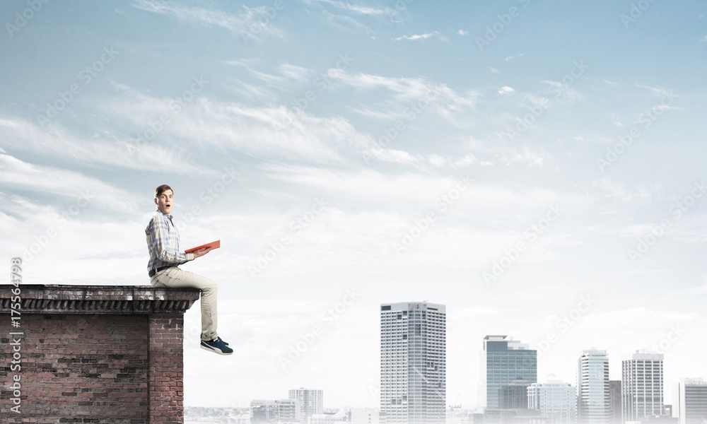 Man on roof edge reading book and cityscape at background Man on roof edge reading book and cityscape at background