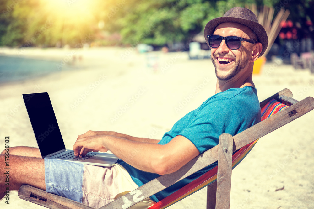 Attractive young man with laptop working on the beach. Freedom, remote work, freelancer, technology, internet, travel and vacation concepts