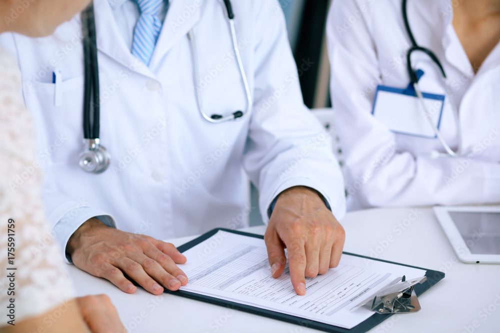 Doctor and patient talking while sitting at the desk in office. Physician pointing into medical history form. Medicine and health care concept Doctor and patient talking while sitting at the desk in office. Physician pointing into medical history form. Medicine and health care concept