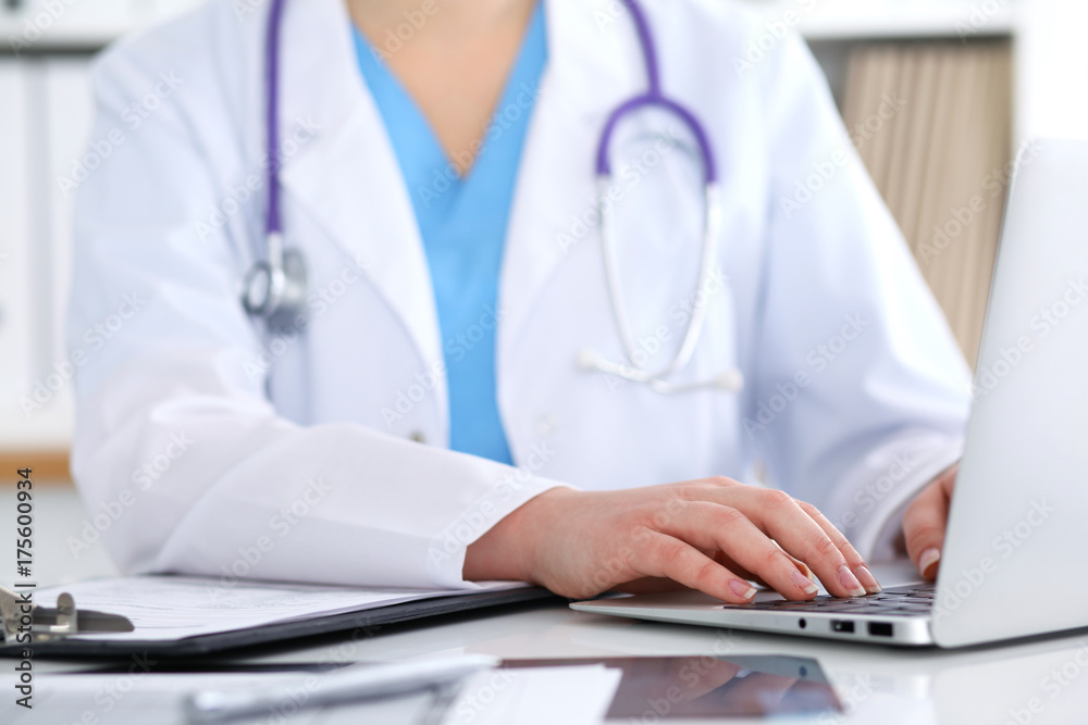 Close up of unknown female doctor typing on laptop computer while sitting  at the table