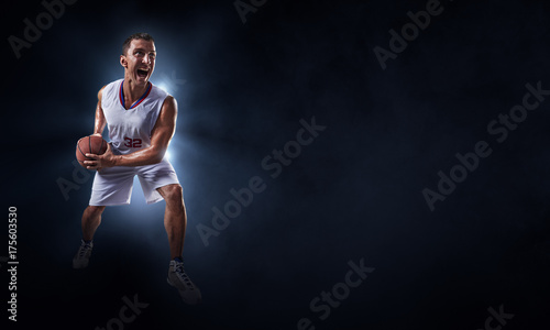 Basketball player stands in the rays of light on a dark background. Player holds a professional basketball ball. Player wears unbranded uniform. © Alex