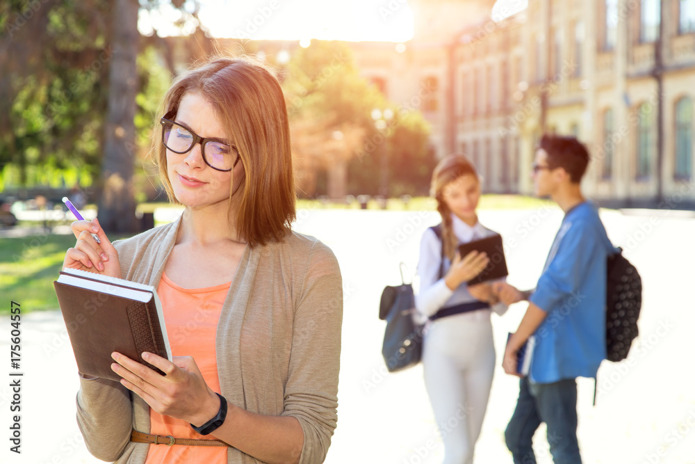 Young female student holds textbook standing in the background of a group of students on the university campus. Student life. Young female student holds textbook standing in the background of a group of students on the university campus. Student life.