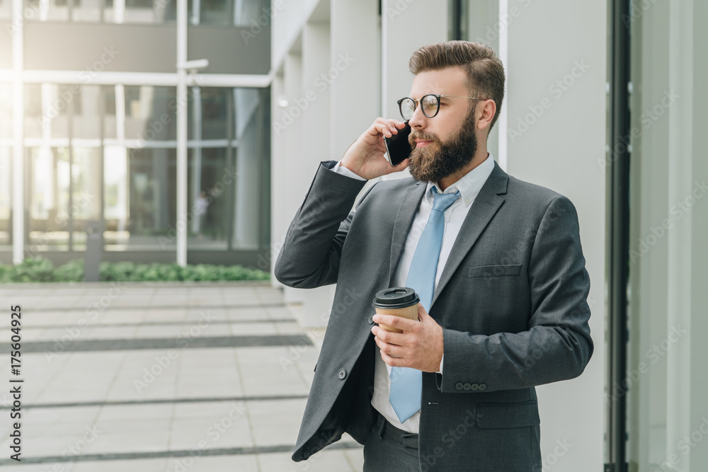 Young businessman in suit and tie is standing outdoor, drinking coffee and talking on his cell phone. In background is modern glass building. Man is working. Negotiations by phone. Business planning. Young businessman in suit and tie is standing outdoor, drinking coffee and talking on his cell phone. In background is modern glass building. Man is working. Negotiations by phone. Business planning.