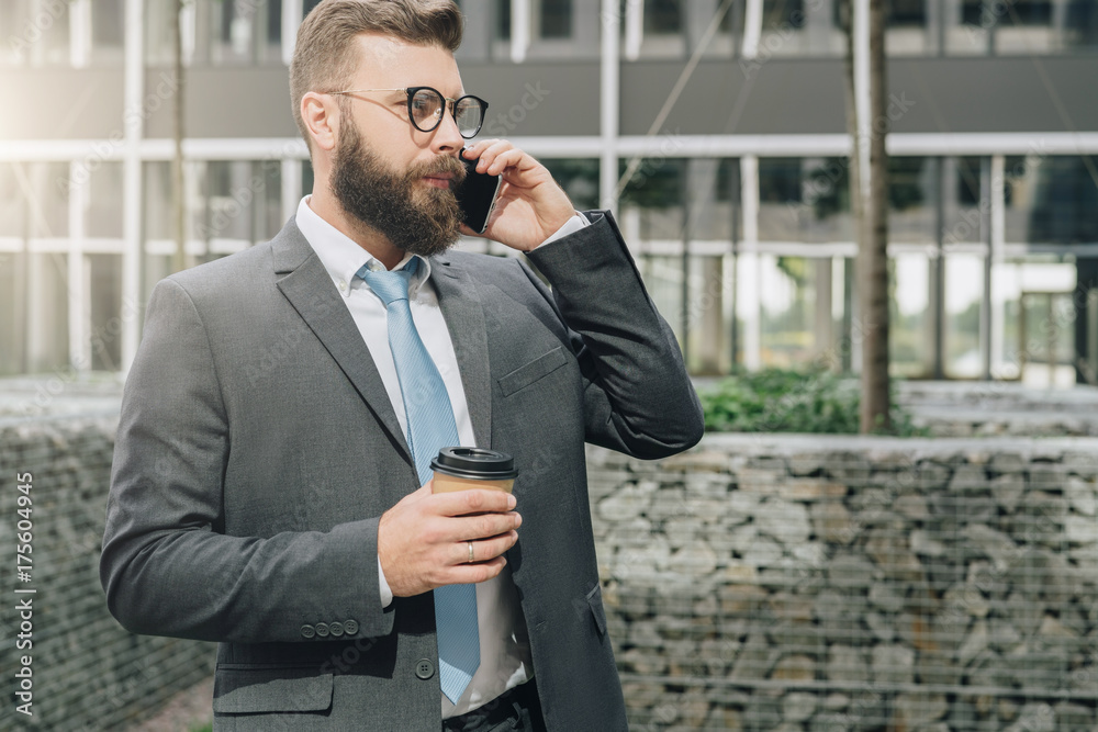 Young businessman in suit and tie is standing outdoor, drinking coffee and talking on his cell phone. In background is modern glass building. Man is working. Negotiations by phone. Business planning. Young businessman in suit and tie is standing outdoor, drinking coffee and talking on his cell phone. In background is modern glass building. Man is working. Negotiations by phone. Business planning.