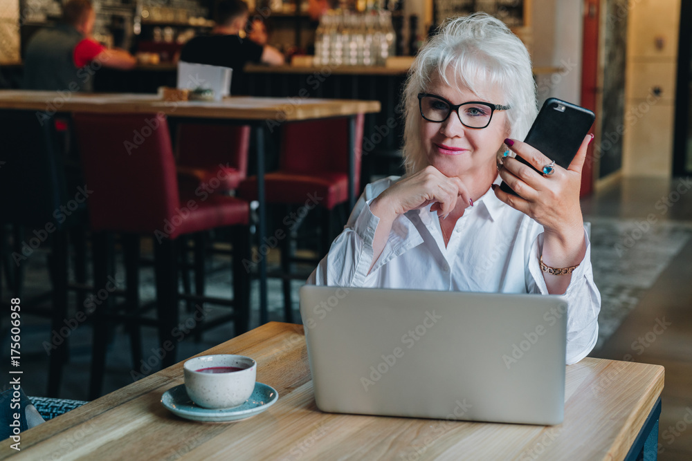 Businesswoman in glasses is sitting at table in front of laptop and holding smartphone.Education for adults. Freelancer works. Retired woman is chatting, blogging, checking email.Social media, network Businesswoman in glasses is sitting at table in front of laptop and holding smartphone.Education for adults. Freelancer works. Retired woman is chatting, blogging, checking email.Social media, network
