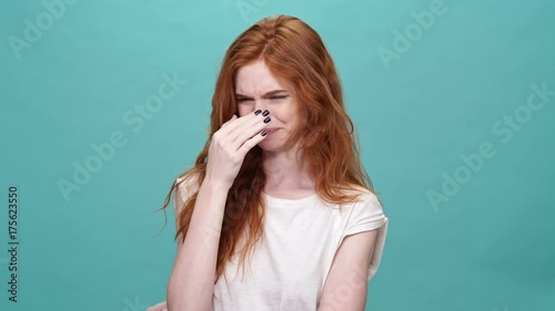 Pretty ginger woman in t-shirt covering her nose because of unpleasant odor over turquoise background