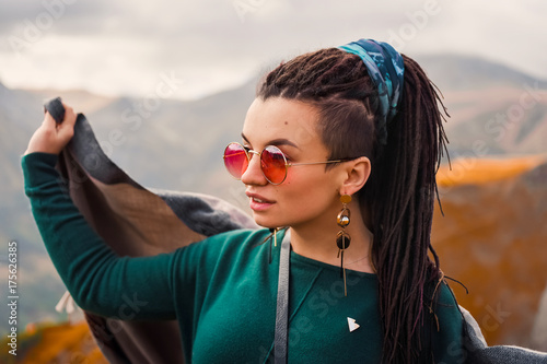 portrait of a young beautiful hipster girl in sunglasses with hair dreadlock in a boho gypsy hippie style clothes on a background of cloudy sky and autumn mountains of Georgia dancing