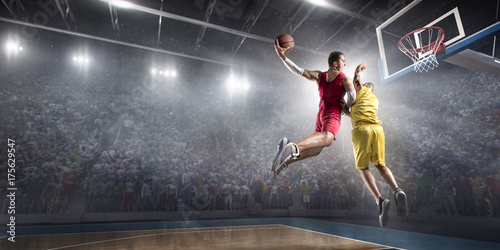 Basketball player makes slam dunk on big professional arena. Player flies through the air with the ball. Opponents try to prevent the ball from hitting the basketball ring.