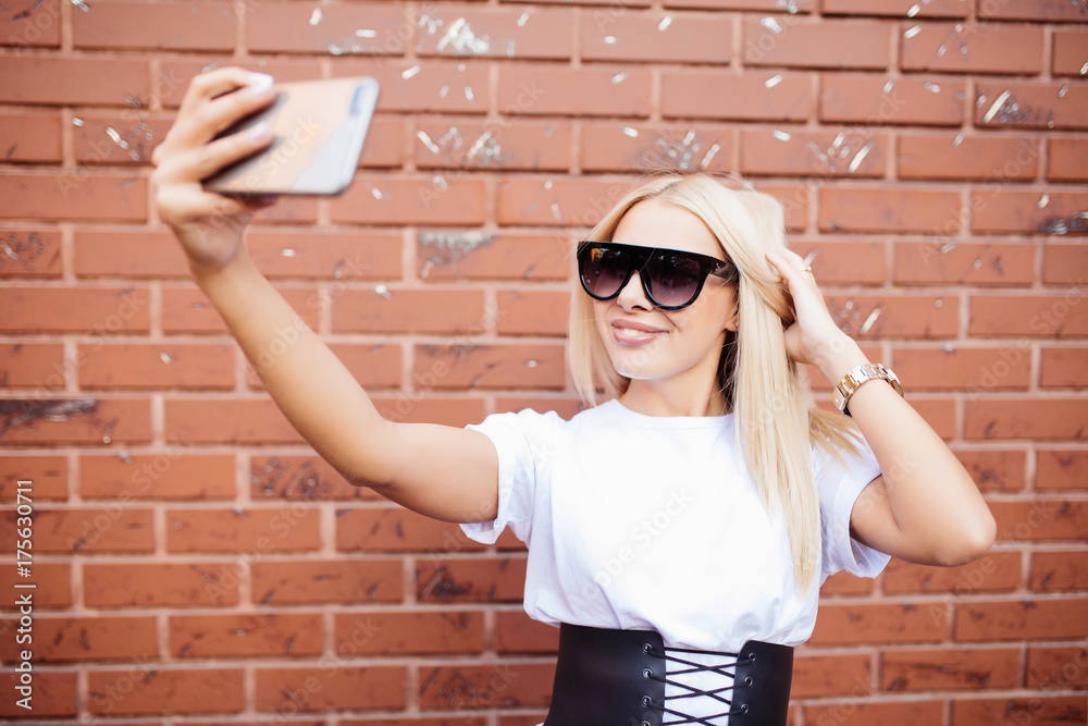 Beautiful blonde woman girl taking a selfie on smartphone, posing standing against red brick wall. Beautiful blonde woman girl taking a selfie on smartphone, posing standing against red brick wall.