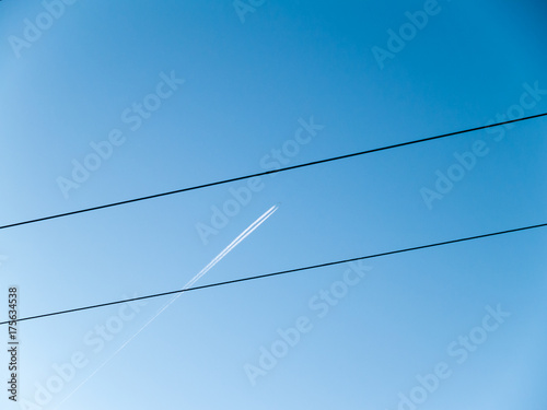 In the clear blue sky, a high flying airplane leaves condensation trail behind itself. The sky is crossed by a line of wires.