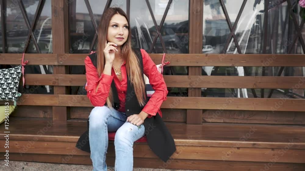 Young beautiful girl in casual clothes sitting on on a wooden bench with cushions in a city park. Young beautiful girl in casual clothes sitting on on a wooden bench with cushions in a city park.