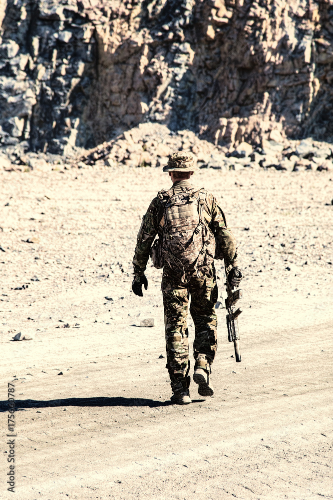 Location shot of soldier in field uniforms with rifle moving in the desert among rocks. Back view