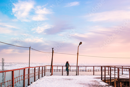 The girl stands in winter on a snow-covered pier leaving in the icy sea against the background of a bright sunset sky.