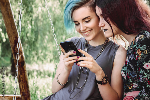 Two girls stits on wooden swing, looking at smartphone. Browsing internet, posting photos in social media photo