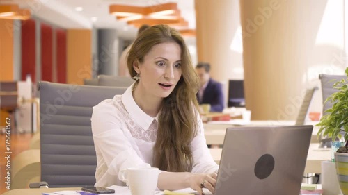 young happy woman shouts and laughs when see good news of success in computer during working day with papers sitting at table in co working colorful business office space