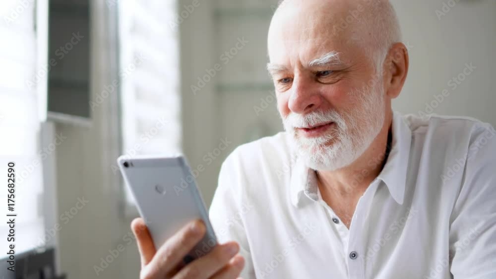 Senior man sitting at home with smartphone. Using mobile talking via messenger app Skype. Smiling waving hand in greeting. Remote communication with children on retirement, active modern lifestyle Senior man sitting at home with smartphone. Using mobile talking via messenger app Skype. Smiling waving hand in greeting. Remote communication with children on retirement, active modern lifestyle