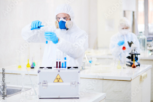 Male researcher wearing hazmat suit adding blue solution to test tube while conducting experiment at modern laboratory